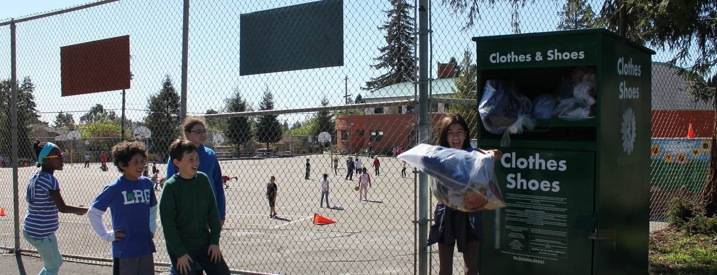 A student laughs as she lifts a bag of clothing into a green collection box beside a school playground, with classmates watching.