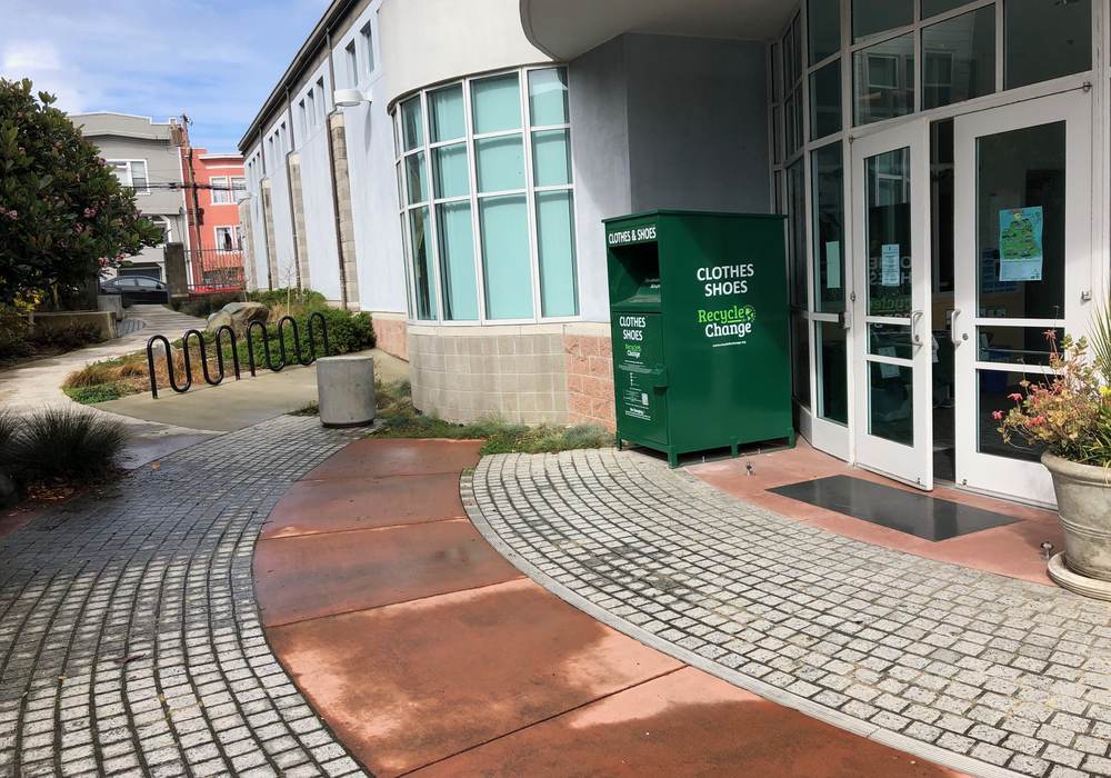 A green Recycle for Change clothes and shoes box standing beside the entrance of a community building.