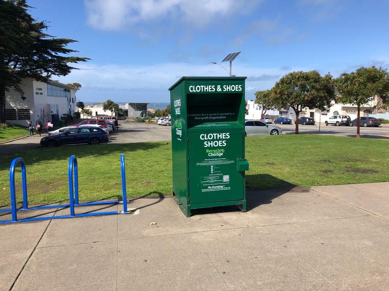 A green Recycle for Change clothes and shoes collection box standing on a grassy verge beside a school, with the ocean behind.