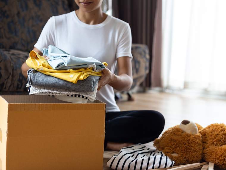 A woman kneels on a living room floor, placing folded clothes into a cardboard donation box.