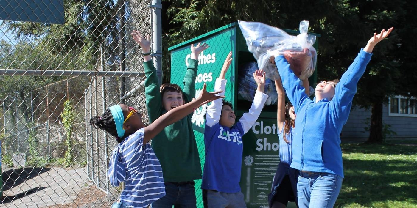 Five schoolchildren throw their arms up as a bag of clothing sails toward a green collection box.