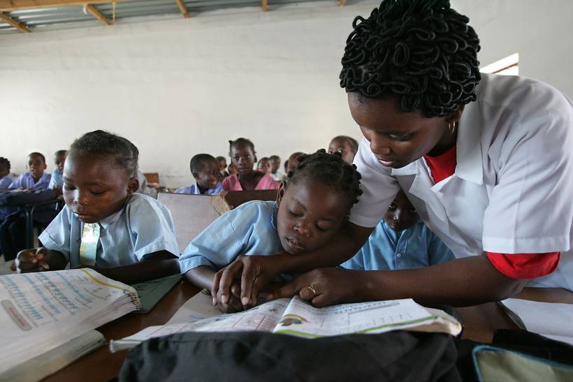 A teacher leans over a desk to guide two young pupils through their exercise books in a busy classroom.