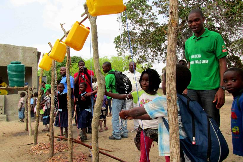 Schoolchildren wash their hands at a row of tippy-tap stations built from wooden poles and suspended jerrycans.