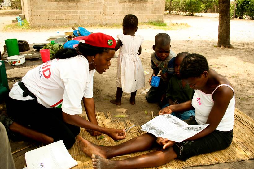 A community health worker sits on a reed mat with a mother, going through an illustrated booklet together while children gather around.