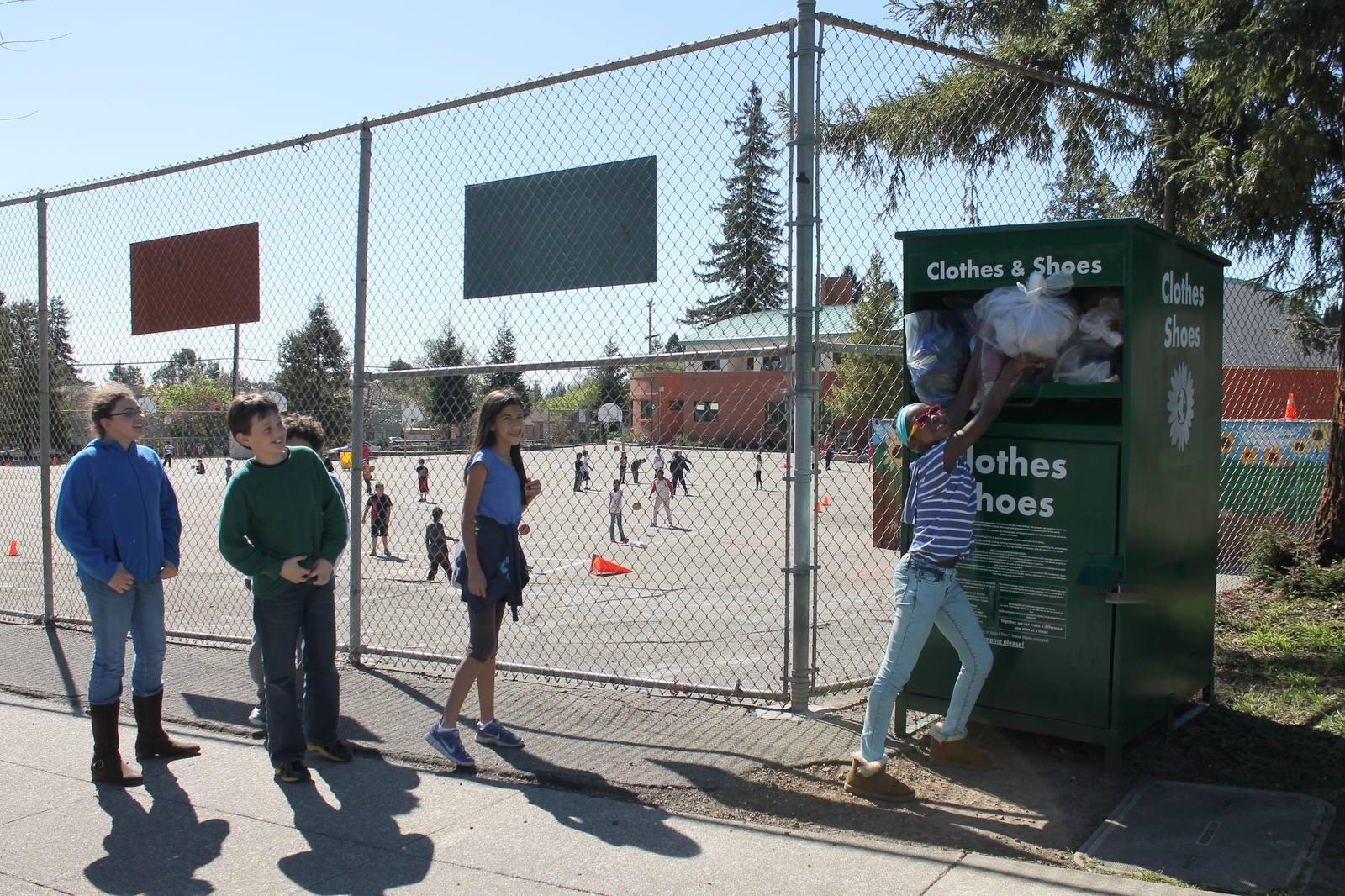 Schoolchildren beside a green Recycle for Change collection box, one lifting a bag of clothing into the chute