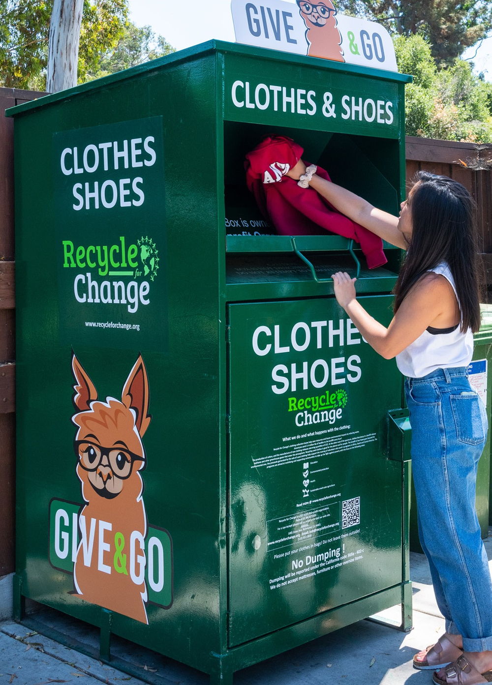 A woman posts a folded red garment through the chute of a green Recycle for Change clothes and shoes collection box, customized with Give and Go branding.
