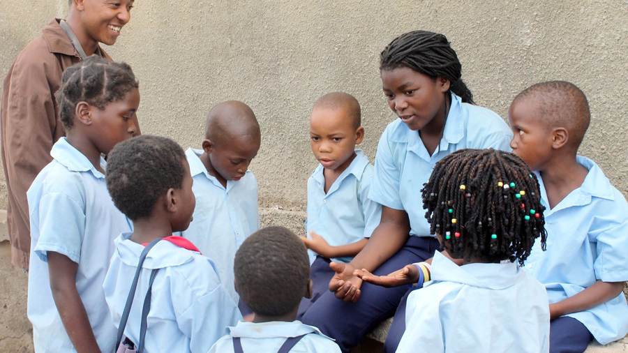 A young woman in school uniform sits talking with a group of primary school children gathered around her.