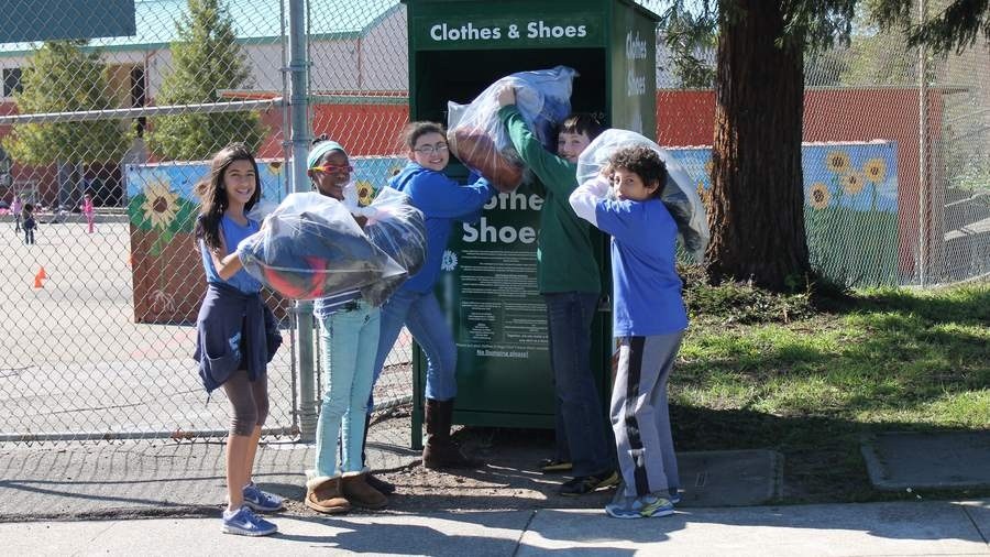 Five schoolchildren smile as they lift bags of clothing into a green Clothes and Shoes collection box in a schoolyard.
