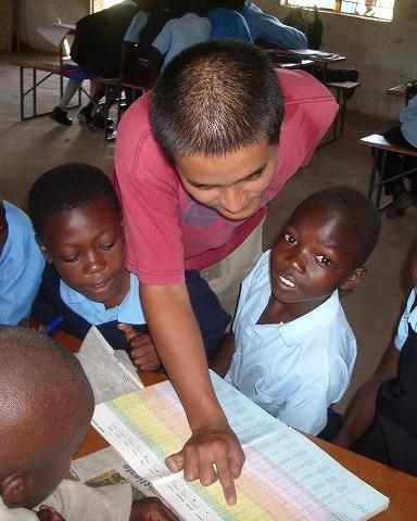 A volunteer leans over a school desk, working through an exercise book with a group of uniformed pupils.