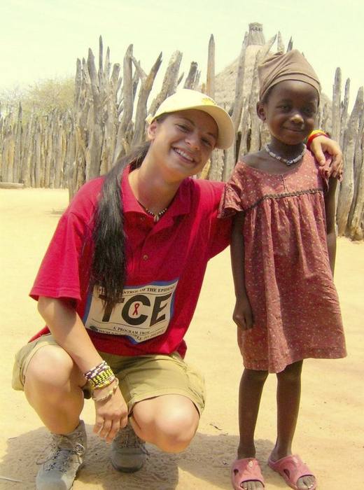 A volunteer in a programme t-shirt crouches beside a young girl outside a village home, both smiling at the camera.