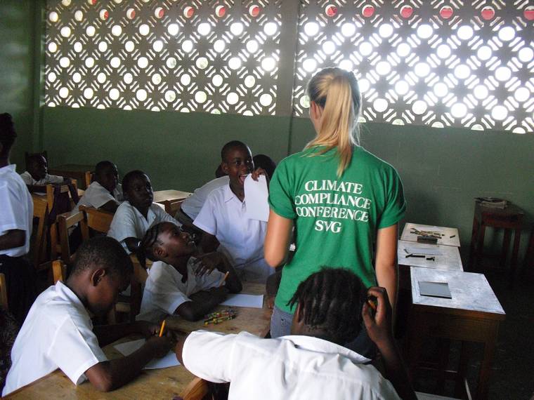A volunteer in a Climate Compliance Conference t-shirt works with pupils at their desks in a classroom.