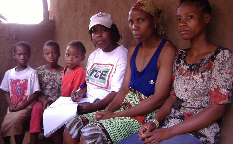 A programme volunteer sits with a family, taking notes in a workbook during a household visit.