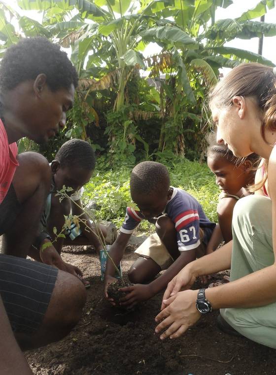 A volunteer and a local trainer plant a seedling with three children in a garden bed.